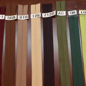 May include: A close-up of a variety of zipper tapes in different colors, including burgundy, brown, beige, cream, black, green, and light blue. The zipper tapes are labeled with numbers and letters, such as "WI", "021", "568", "810", "125", "Z122", "AG", "GN", "LGN", and "544".