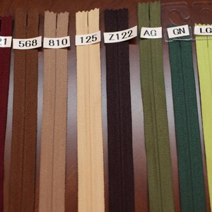 Puede incluir: Primer plano de una variedad de cintas de cremallera en diferentes colores, incluyendo burdeos, marr&oacute;n, beige, crema, negro, verde y azul claro. Las cintas de cremallera est&aacute;n etiquetadas con n&uacute;meros y letras, como "WI", "021", "568", "810", "125", "Z122", "AG", "GN", "LGN" y "544".