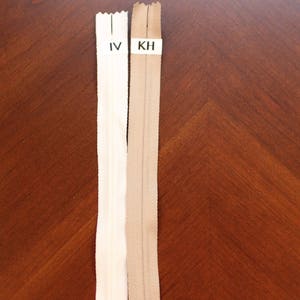 May include: Two zipper tapes, one white and one beige, are laid out on a wooden surface. The white zipper tape has the label "IV" and the beige zipper tape has the label "KH".