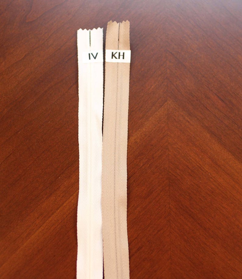 May include: Two zipper tapes, one white and one beige, are laid out on a wooden surface. The white zipper tape has the label "IV" and the beige zipper tape has the label "KH".