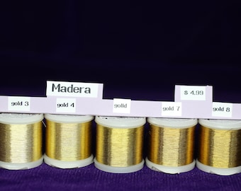 Könnte beinhalten: Fünf Spulen goldfarbener Madeira-Garn, beschriftet mit "gold 3", "gold 4", "gold", "gold 7" und "gold 8". Die Spulen sind in einer Reihe auf einer violetten Oberfläche angeordnet. Ein Preisschild zeigt "4,99 €".