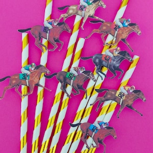 May include: A collection of paper straws with gold and white stripes, arranged diagonally against a bright pink background. Small cutouts of racehorses with jockeys are attached to the straws, creating a festive theme.