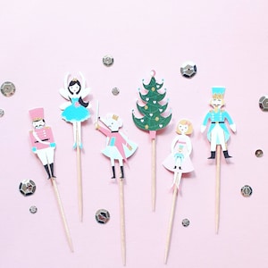 May include: A collection of festive cupcake toppers featuring a pink and blue color scheme. The toppers include a ballerina, a Christmas tree, a mouse, a soldier, and a nutcracker. The toppers are on wooden sticks, perfect for holiday celebrations.