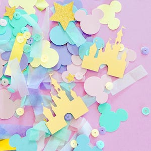 May include: A pastel-colored confetti arrangement on a pink background. The confetti includes Mickey Mouse silhouettes, castle cutouts, stars, and sequins in shades of yellow, blue, purple, and pink. There are also strips of translucent paper.