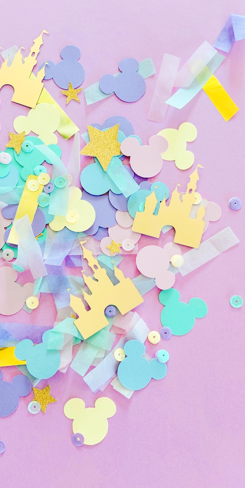 May include: A pastel-colored confetti arrangement on a pink background. The confetti includes Mickey Mouse silhouettes, castle cutouts, stars, and sequins in shades of yellow, blue, purple, and pink. There are also strips of translucent paper.