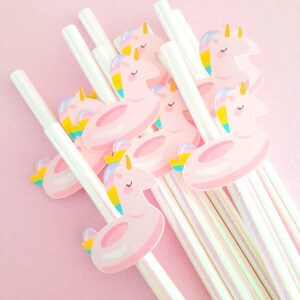 May include: A set of iridescent white striped straws with unicorn-shaped toppers. Each topper features a pink inflatable ring and a unicorn with a rainbow mane and tail.