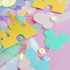 May include: A collection of pastel-colored paper cutouts, including Mickey Mouse silhouettes, castle shapes, stars, and cloud shapes, scattered on a pink background. The cutouts are in shades of yellow, blue, pink, and purple, with some glitter accents.