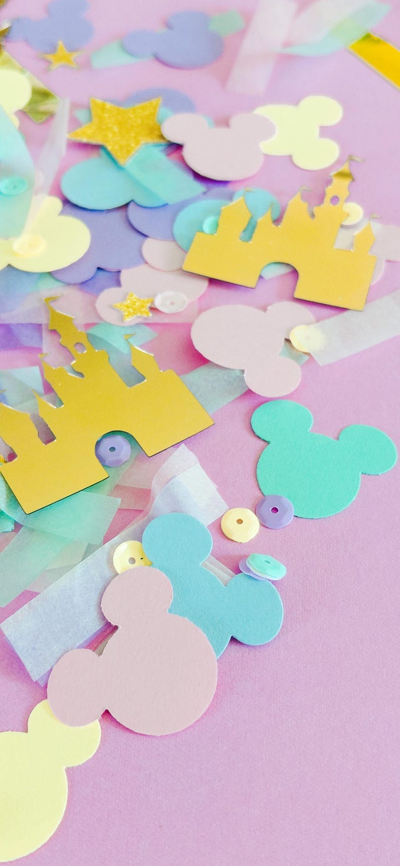 May include: A collection of pastel-colored paper cutouts, including Mickey Mouse silhouettes, castle shapes, stars, and cloud shapes, scattered on a pink background. The cutouts are in shades of yellow, blue, pink, and purple, with some glitter accents.