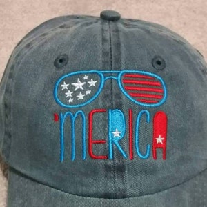 Op de afbeelding: Een blauwe denim baseballcap met een geborduurd ontwerp van een zonnebril met een Amerikaans vlagpatroon en het woord "'MERICA" in rood en blauw garen.