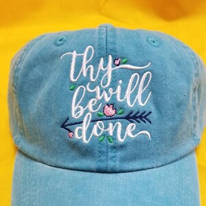 May include: A light blue baseball cap with white embroidery that reads "thy will be done". The embroidery includes a small pink flower and an arrow.