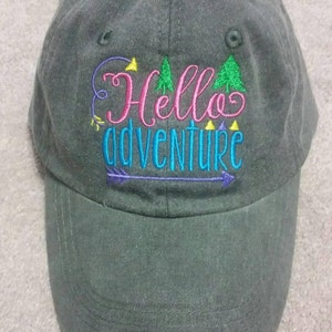 May include: A dark green baseball cap with the embroidered text "Hello Adventure" in pink, teal, and purple. The design includes trees and an arrow. The cap has a curved brim and a button on top.