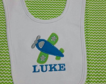 May include: White bib with a green airplane and stars design. The bib is embroidered with the name "LUKE".