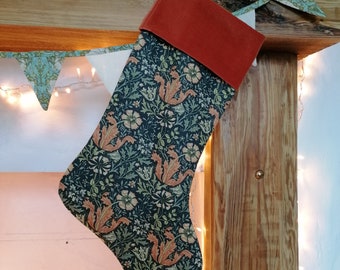 May include: A Christmas stocking with a dark teal floral pattern and a rust-colored velvet cuff. The fabric features a repeating design of stylized flowers and leaves in shades of orange, green, and cream. The stocking is hanging on a wooden mantle.