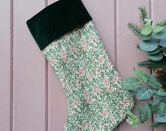 May include: A Christmas stocking with a green velvet cuff and a floral pattern. The pattern features green leaves and pink flowers on a cream background. The stocking is hanging on a pink wooden surface.
