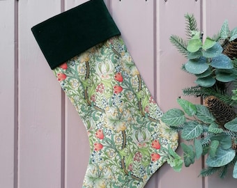 May include: A Christmas stocking with a green velvet cuff and a floral pattern in shades of green, yellow, and red. The stocking is hanging on a pink wooden surface. A decorative wreath is in the background.