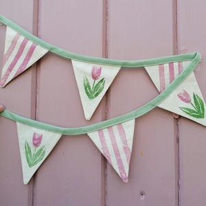 May include: A decorative banner with triangular flags. The flags are cream-colored with pink stripes and tulip designs. The banner is trimmed with a green border and is hanging against a pink background.