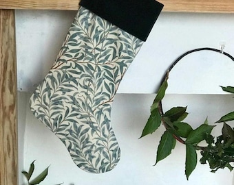 May include: A Christmas stocking with a black cuff and a cream-colored body featuring a blue and green leaf pattern. The stocking is hanging on a white wall, with a wooden shelf above it. A mug with a bird design is on the shelf.