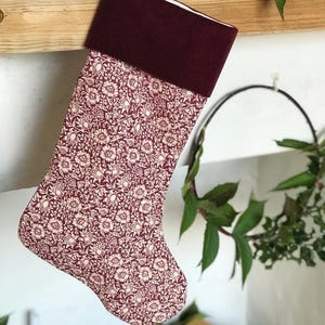 Op de afbeelding: Een kerstsok met een bordeauxrode manchet en een bloemenpatroon in bordeauxrood en crème. De sok hangt aan een houten schoorsteenmantel. De achtergrond omvat groen en een ingepakt cadeau.