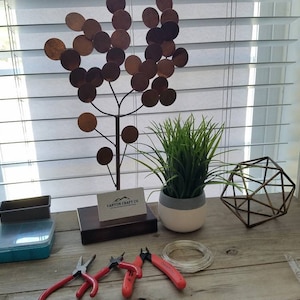 May include: A rustic wooden table displays craft supplies and decorative items. A metal tree sculpture with circular leaves stands near a potted plant and a geometric wireframe. Red-handled pliers and wire spools are also visible.
