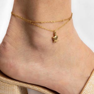 May include: A double-layered gold anklet featuring a delicate chain with small beads and a central turtle charm. The piece is crafted from a polished gold-tone metal, showcasing a minimalist design with a small, detailed sea turtle pendant.