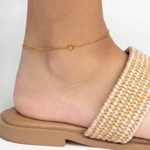 May include: A gold-toned anklet featuring a delicate chain interspersed with small, open heart-shaped charms. The jewelry piece rests around the ankle, showcasing a simple link design that connects each heart element in a continuous, uniform pattern.