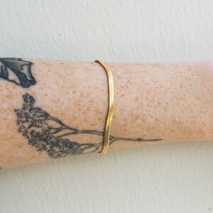 Puede incluir: Un brazalete de cadena dorada en el brazo de una persona con un tatuaje de flores.