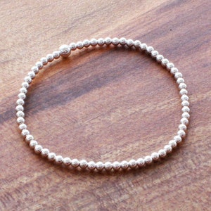 以下が含まれることがあります： シンプルなデザインのシルバービーズブレスレット。ブレスレットは、細いワイヤーに小さな丸いビーズが連なって作られています。ブレスレットは木製の表面に平らに置かれています。