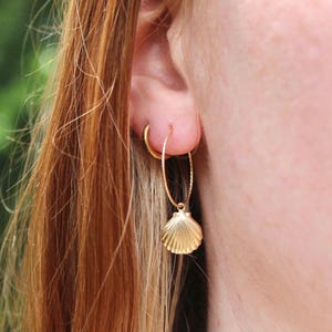 May include: A close-up of a gold hoop earring with a textured design, and a small gold seashell charm. The earring is worn on a person's ear, with their hair visible in the background. The seashell charm is a detailed, miniature replica.