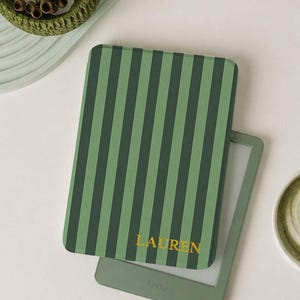 May include: Green Kindle with a cover featuring vertical stripes in shades of green. The cover has the name "LAUREN" in gold lettering. A cup of matcha latte and decorative items are also in the image.