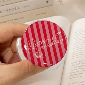 May include: A circular phone grip featuring a red and pink vertical striped pattern. The text "Rather be reading" is printed in white script across the center, accompanied by a small white line-art illustration of an open book and two hearts.