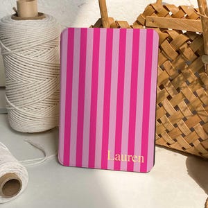 May include: A pink and light pink striped notebook with the name "Lauren" in gold lettering. The notebook is rectangular and stands upright. A roll of white twine and a woven basket are in the background.