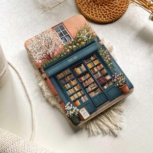 Op de afbeelding: Een ge&iuml;llustreerde roze en blauwe boekwinkel met een bord waarop "Book Shop" staat. De winkel heeft een etalage met boeken en een bloempot met witte bloemen. De afbeelding is op een witte achtergrond.