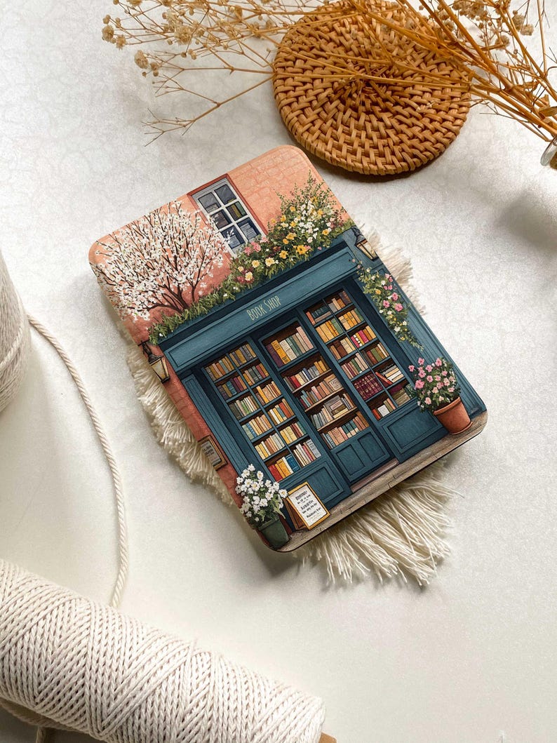 Op de afbeelding: Een ge&iuml;llustreerde roze en blauwe boekwinkel met een bord waarop "Book Shop" staat. De winkel heeft een etalage met boeken en een bloempot met witte bloemen. De afbeelding is op een witte achtergrond.