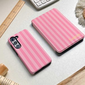 以下が含まれることがあります： ピンクのストライプ柄の携帯電話ケース2つと、金色の文字で「Lauren」と書かれたお揃いのケース。電卓と木製の定規も白い表面に見えます。