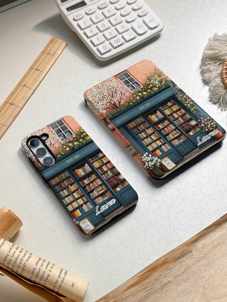 Op de afbeelding: Twee telefoonhoesjes met een boekwinkelontwerp. De hoesjes zijn beide roze en blauw met een boekenkast vol boeken. De tekst "BOOK SHOP" staat op de voorgevel van de winkel. De naam "Lauren" staat op elk hoesje.