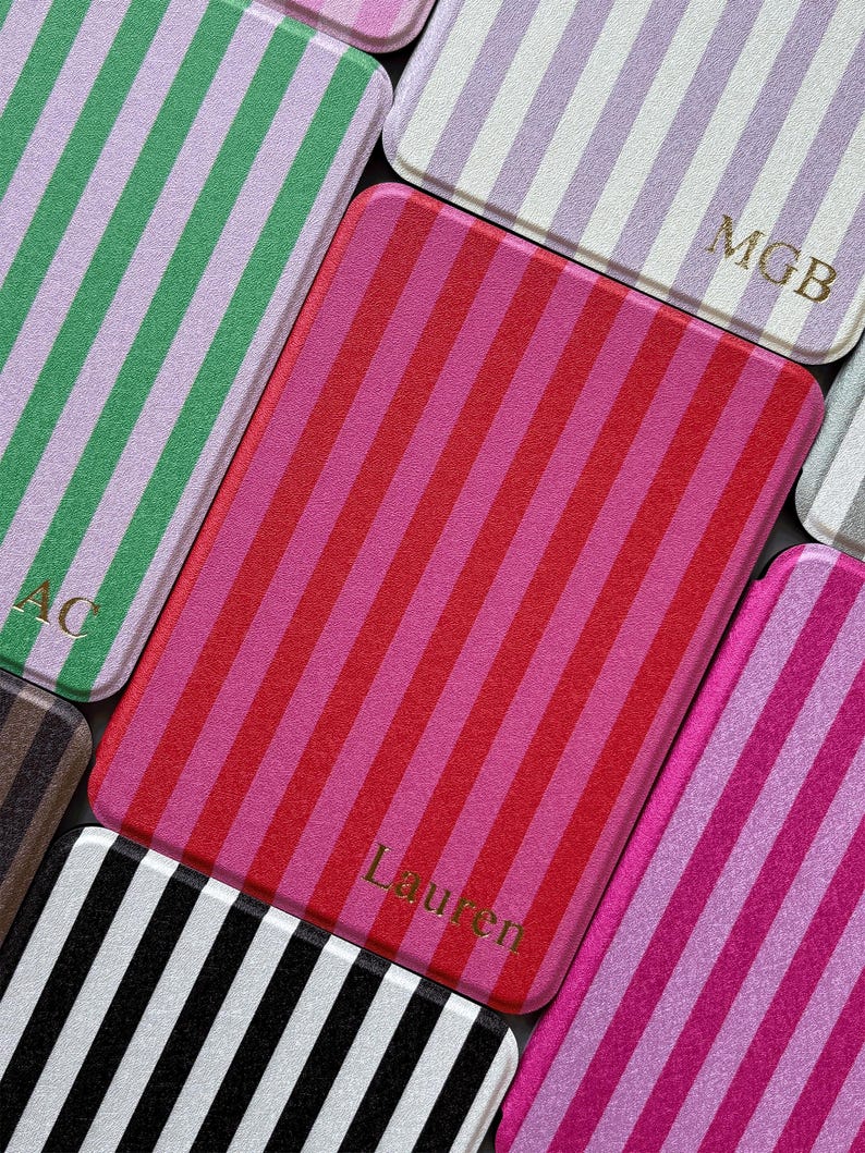 May include: Close-up of several rectangular cases with vertical stripes in various colours, including pink, red, green, and black. Some cases have gold lettering with initials or a name. The cases have rounded corners.