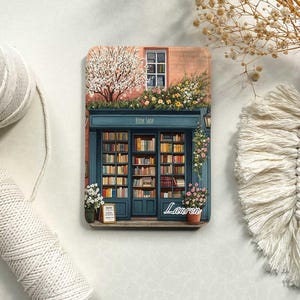 Op de afbeelding: Een kleurrijke illustratie van een boekwinkel met een bord waarop "Book Shop" staat. De winkel heeft een groot raam met planken vol boeken. Er zijn bloemen en een boom voor de winkel. De naam "Lauren" staat onder de winkel.
