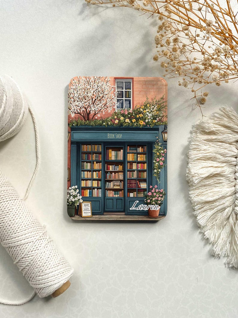Op de afbeelding: Een kleurrijke illustratie van een boekwinkel met een bord waarop "Book Shop" staat. De winkel heeft een groot raam met planken vol boeken. Er zijn bloemen en een boom voor de winkel. De naam "Lauren" staat onder de winkel.