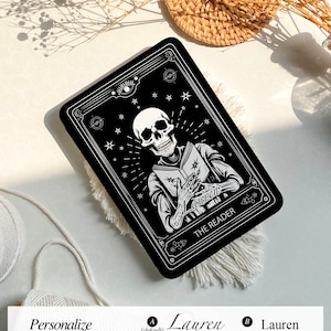 Puede incluir: Una ilustración en blanco y negro de un esqueleto leyendo un libro con el texto "THE READER" sobre un fondo negro. La ilustración está rodeada por un borde decorativo con estrellas y otros símbolos.