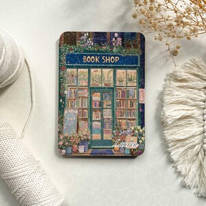May include: A green and blue illustration of a book shop with a sign that reads "BOOK SHOP". The shop has a window display with books and plants. The illustration is set against a snowy background. The name "Lauren" is written in white at the bottom of the illustration.