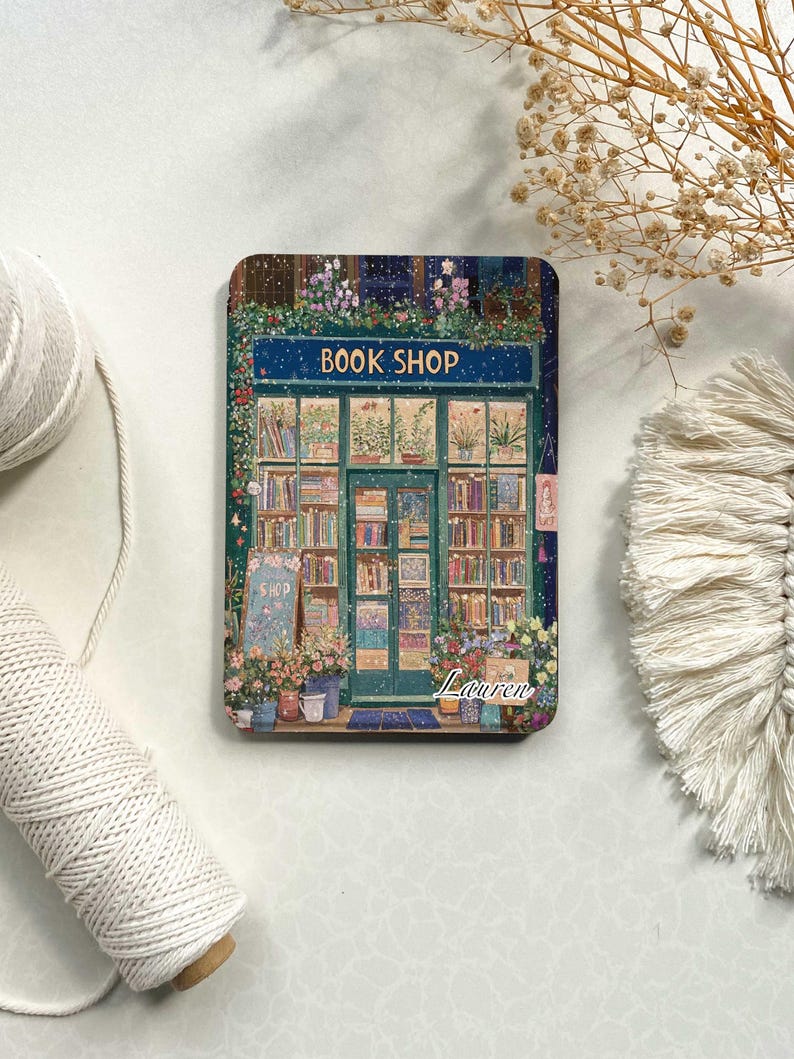 May include: A green and blue illustration of a book shop with a sign that reads "BOOK SHOP". The shop has a window display with books and plants. The illustration is set against a snowy background. The name "Lauren" is written in white at the bottom of the illustration.