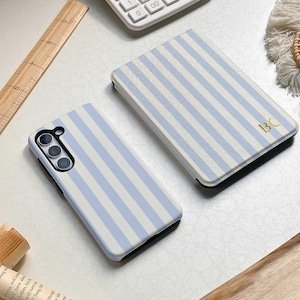 Puede incluir: Dos fundas para teléfono y una funda con un patrón de rayas azul claro y blanco. La funda tiene las iniciales doradas "BC". También se ven una calculadora y una regla de madera.