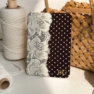 Puede incluir: Un cuaderno rectangular con una cubierta de color marrón oscuro con lunares blancos en el lado derecho y una franja vertical de encaje floral blanco en el izquierdo. La esquina inferior derecha muestra las letras mayúsculas doradas BC.
