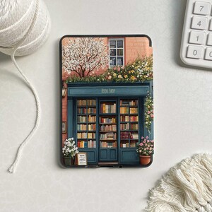 Op de afbeelding: Een zwarte e-reader met een kleurrijke illustratie van een boekwinkel met een bord waarop 'Book Shop' staat. De boekwinkel heeft een raam met uitzicht op een boom met witte bloesems en een bakstenen gebouw met een roze gevel.