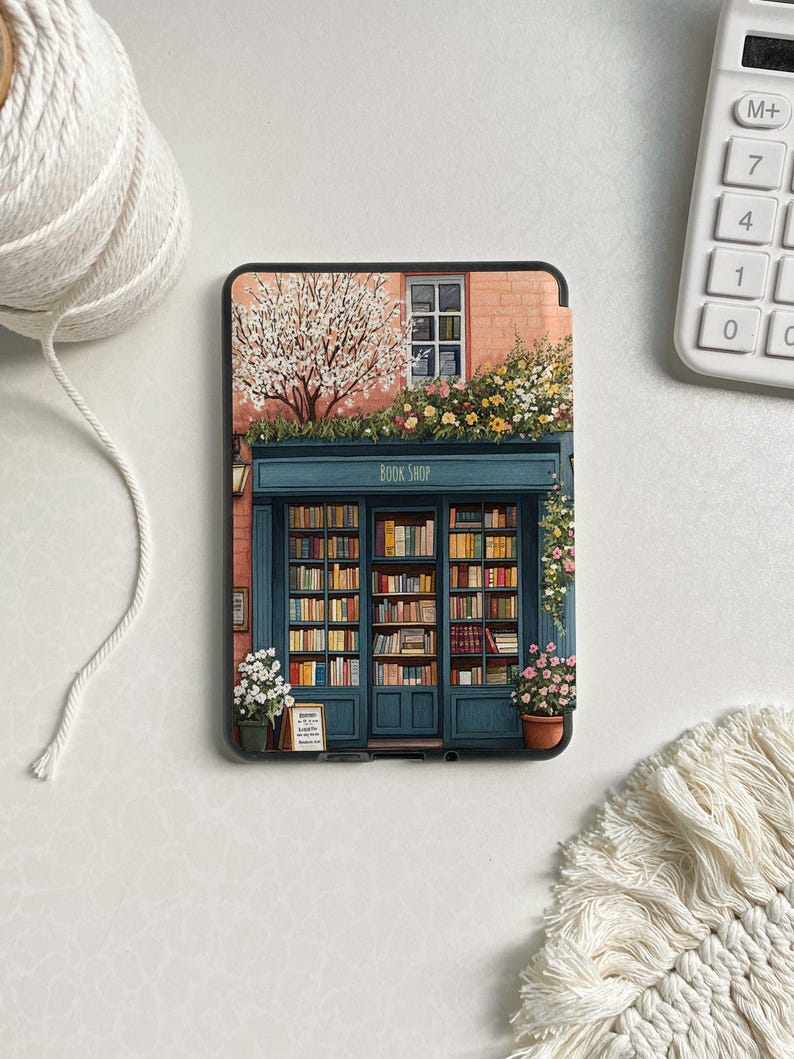 Op de afbeelding: Een zwarte e-reader met een kleurrijke illustratie van een boekwinkel met een bord waarop 'Book Shop' staat. De boekwinkel heeft een raam met uitzicht op een boom met witte bloesems en een bakstenen gebouw met een roze gevel.