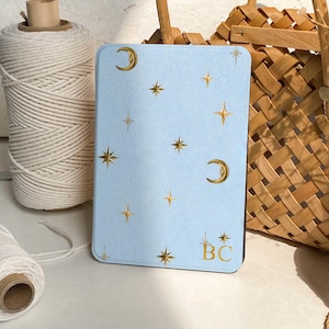 May include: A rectangular light blue tablet or e-reader case featuring a gold foil celestial pattern of crescent moons and various star shapes. The bottom right corner displays the gold foil letters BC, each accented with a small star design.