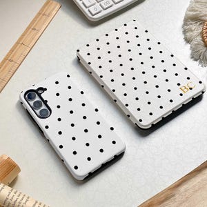 May include: Two white phone cases with black polka dots. One case is lying flat, and the other is angled. A calculator, a wooden ruler, and a book are in the background. The second case has the initials "BC" in gold.