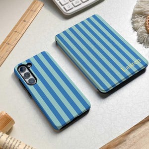 May include: Two phone cases with a blue and light blue striped pattern. One case is open, revealing the phone's camera and buttons. The other case is closed, with the name "Lauren" in gold lettering. A calculator and wooden ruler are in the background.