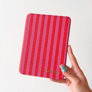 K&ouml;nnte beinhalten: Eine rechteckige E-Reader-H&uuml;lle mit vertikalem Streifenmuster in abwechselnden Rot- und Rosat&ouml;nen. Der Name Lauren ist in goldener Schrift nahe der unteren rechten Ecke aufgedruckt. Der Text style 01 erscheint in der oberen rechten Ecke.