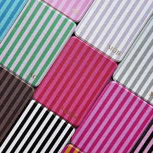 Puede incluir: Una colección de estuches rectangulares con rayas verticales en varios colores, incluyendo rosa, verde, negro y dorado. Algunos estuches tienen iniciales o nombres en letras doradas. Los estuches están dispuestos en diagonal.